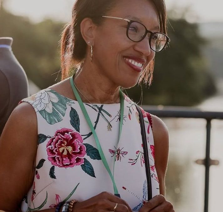 Rachida Presotto, femme souriante qui porte des lunettes noires et une robe avec des motifs à fleurs, bijoux (bracelets, bagues, montres, boucles d'oreilles, coucher de soleil. elle a les cheveux noirs attachés avec une pinces.