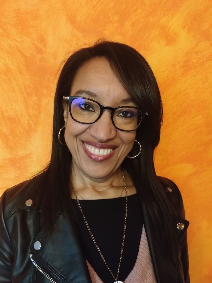 Rachida Presotto, femme souriante qui porte des lunettes noires, un sweat noir, un pull rose, un blouson en simili cuir noir, bijoux (colliers, boucles d'oreilles). elle a les cheveux noirs détachés. le fond de la photo est jaune-orange