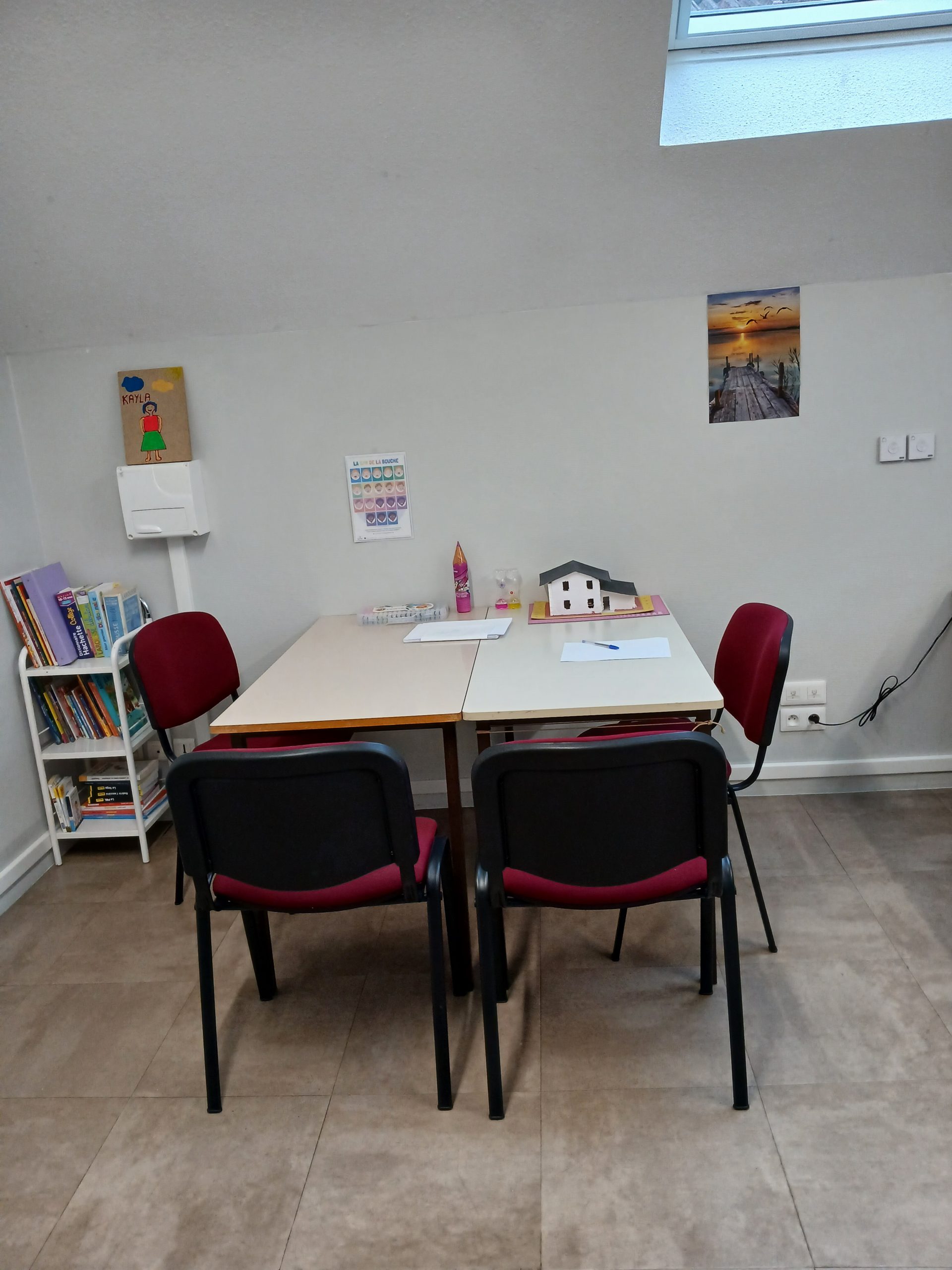 une bibliothèque avec des livres, 2 chaises de couleur bordeau, 2 tables de couleurs blanches avec les pieds marrons, une lampe de chvet/bureau, une boite de feutres, un sablier jaune et rose, une boite de crayons couleurs en forme de crayons, une image de coucher de soleil avec un pont