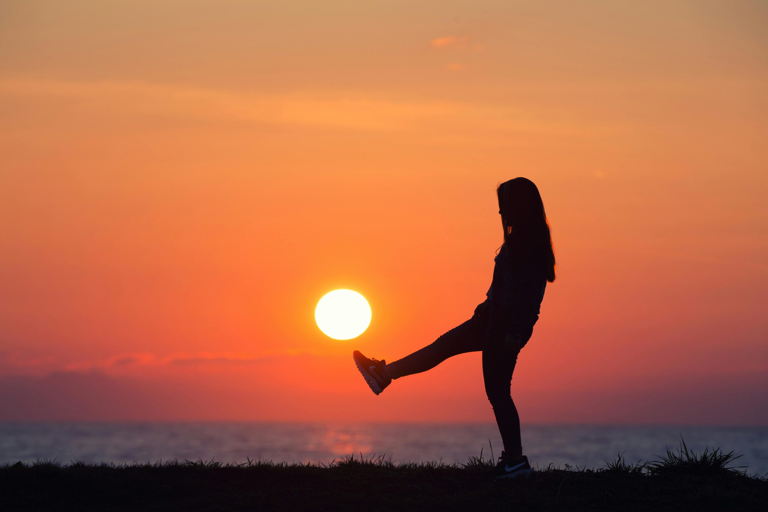 coucher de soleil avec toutes les nuances de couleur (jaune, orange, bleu, gris). l'image est prise en bord de mer. la femme est sur la plage, elle porte des basket rouge et a le pied droit levé comme si elle voulait shooter le soleil.
