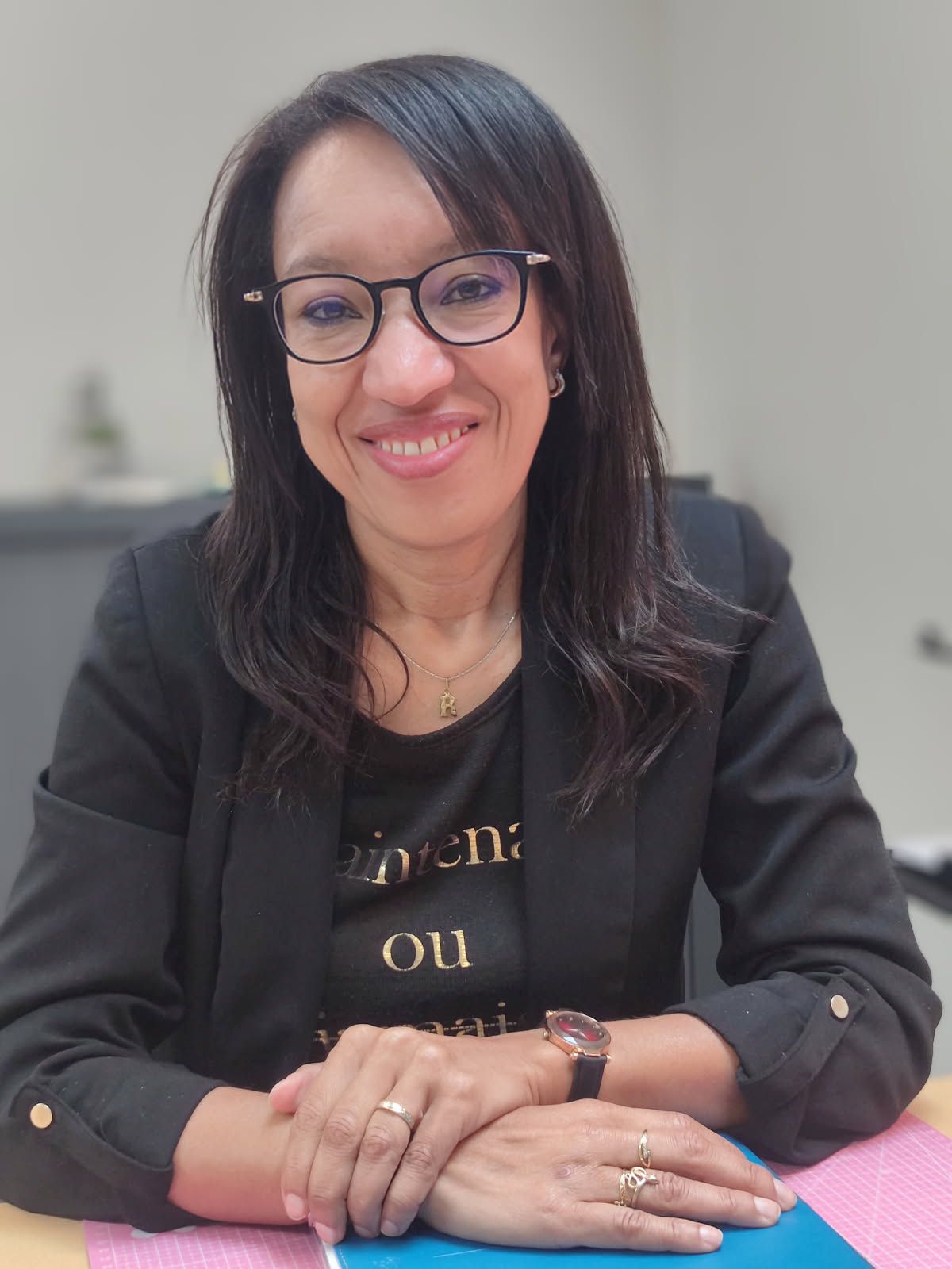 Rachida Presotto, femme souriante qui porte des lunettes noires et une veste noire et tee-shirt noir avec des écritures. Elle a une main posée sur le poignet de l'autre main, elle porte des bijoux (collier avec une lettre R, montre dorée avec bracelet noir, bagues, boucles d'oreilles. Elle est assise et pose devant son bureau. Ses cheveux sont noirs et détachés.