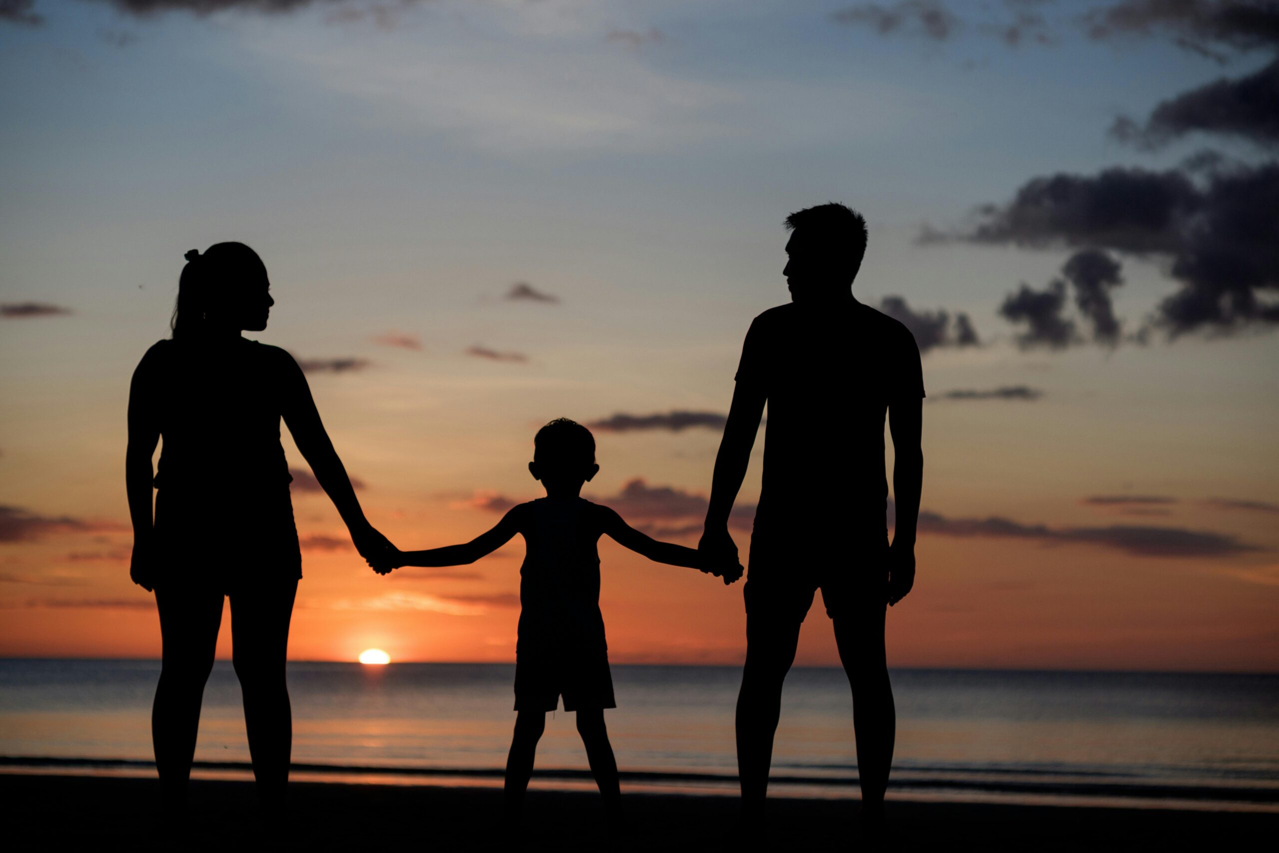 coucher de soleil avec toutes les nuances de couleur (jaune, orange, bleu, gris). l'image est prise en bord de mer. la famille (papa, maman et leur enfant) sont la plage. les parents tiennent la main de leur enfant qui est au milieu.